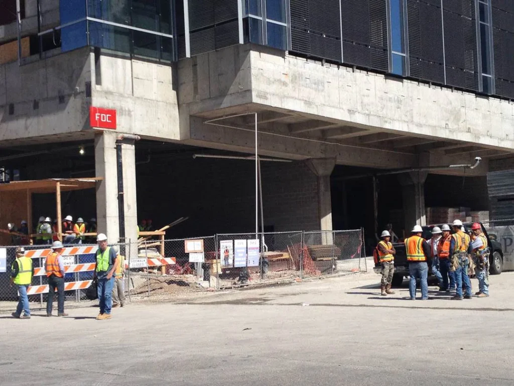 Crews in safety vests at a downtown construction site where barricades and a closed sidewalk meet the public street